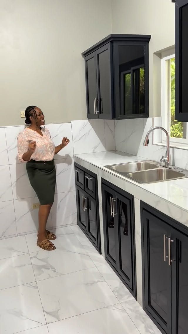Black kitchen cabinets with white marble countertops and floors and a woman standing.