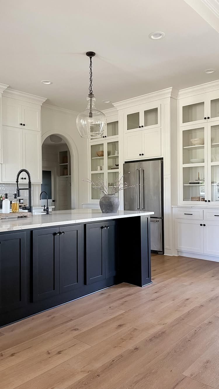 Black kitchen island with white marble top against white cabinets and wood floors