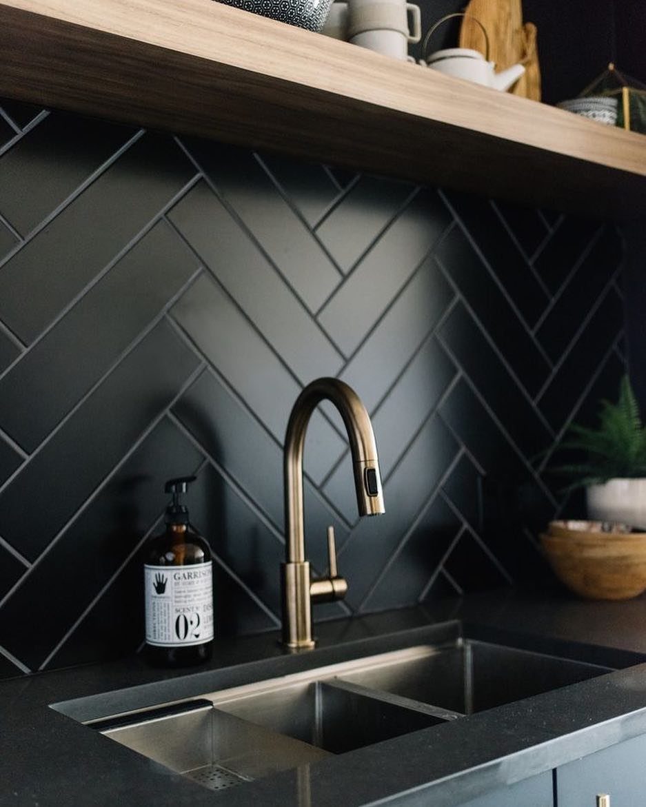 Black herringbone tile backsplash with a gold faucet and light wood floating shelf.