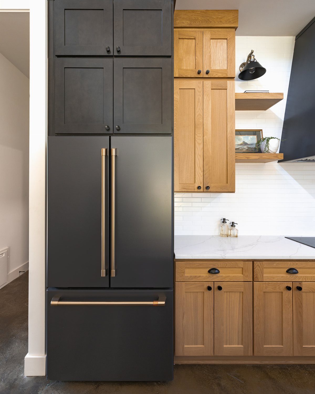 Built-in black refrigerator with gold handles next to light wood kitchen cabinets.