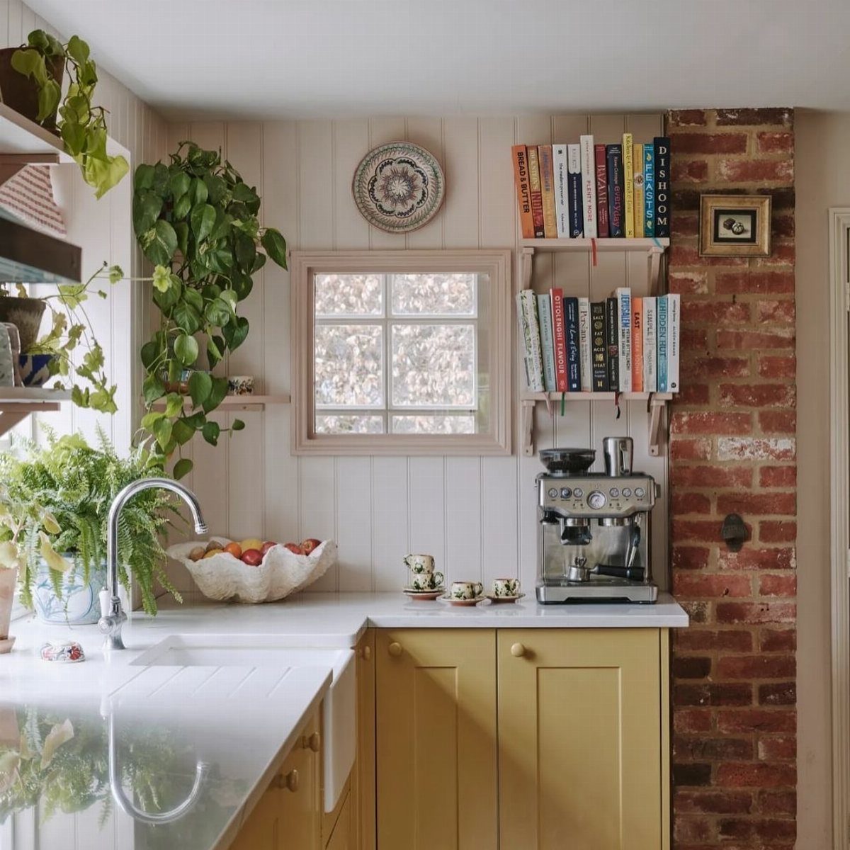 Butter yellow kitchen cabinets with white countertops, a brick wall, and indoor plants.