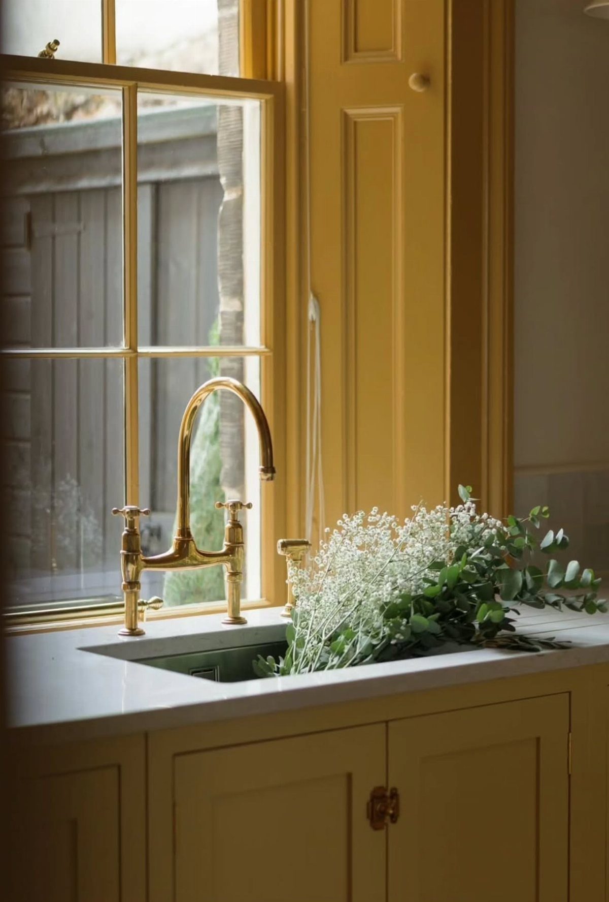 Butter yellow kitchen cabinets and window trim with a gold faucet and white flowers.