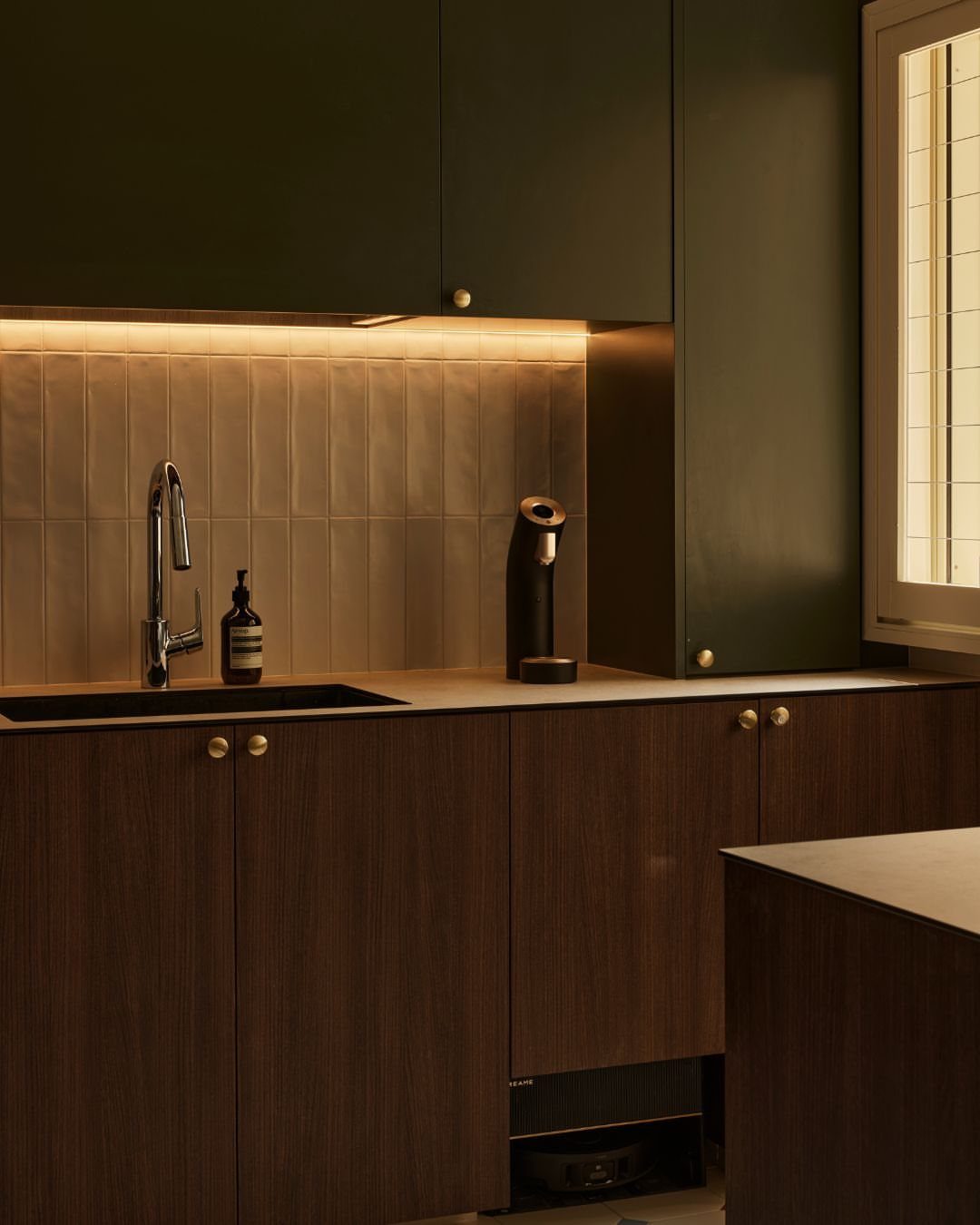 Dark green upper cabinets with warm under-cabinet lighting illuminating a tiled kitchen backsplash.