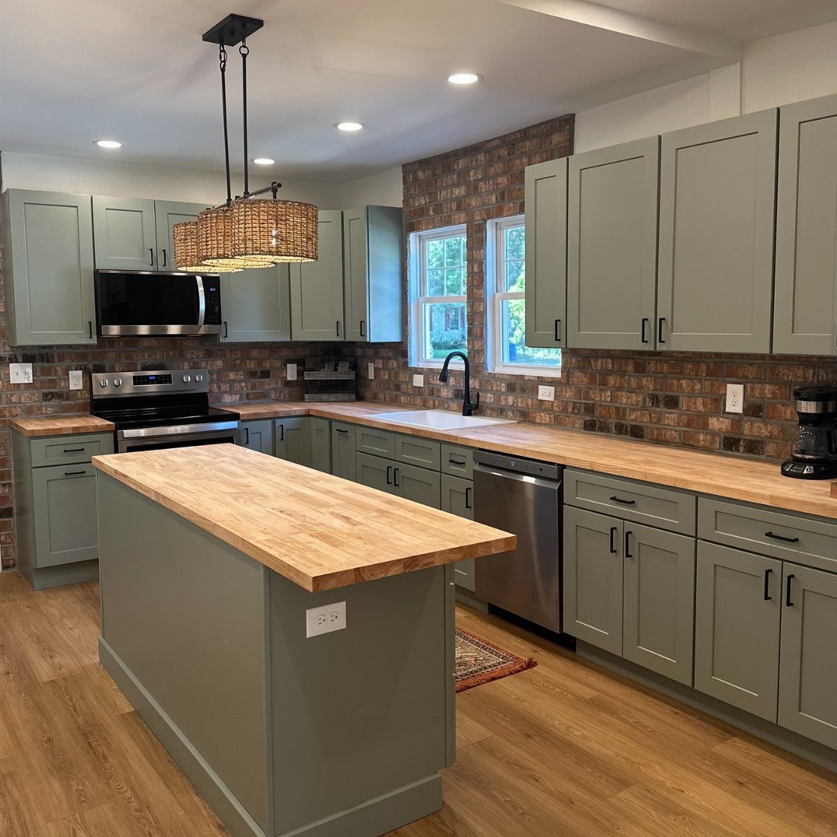 Sage green kitchen cabinets with light wood countertops and a red brick backsplash.