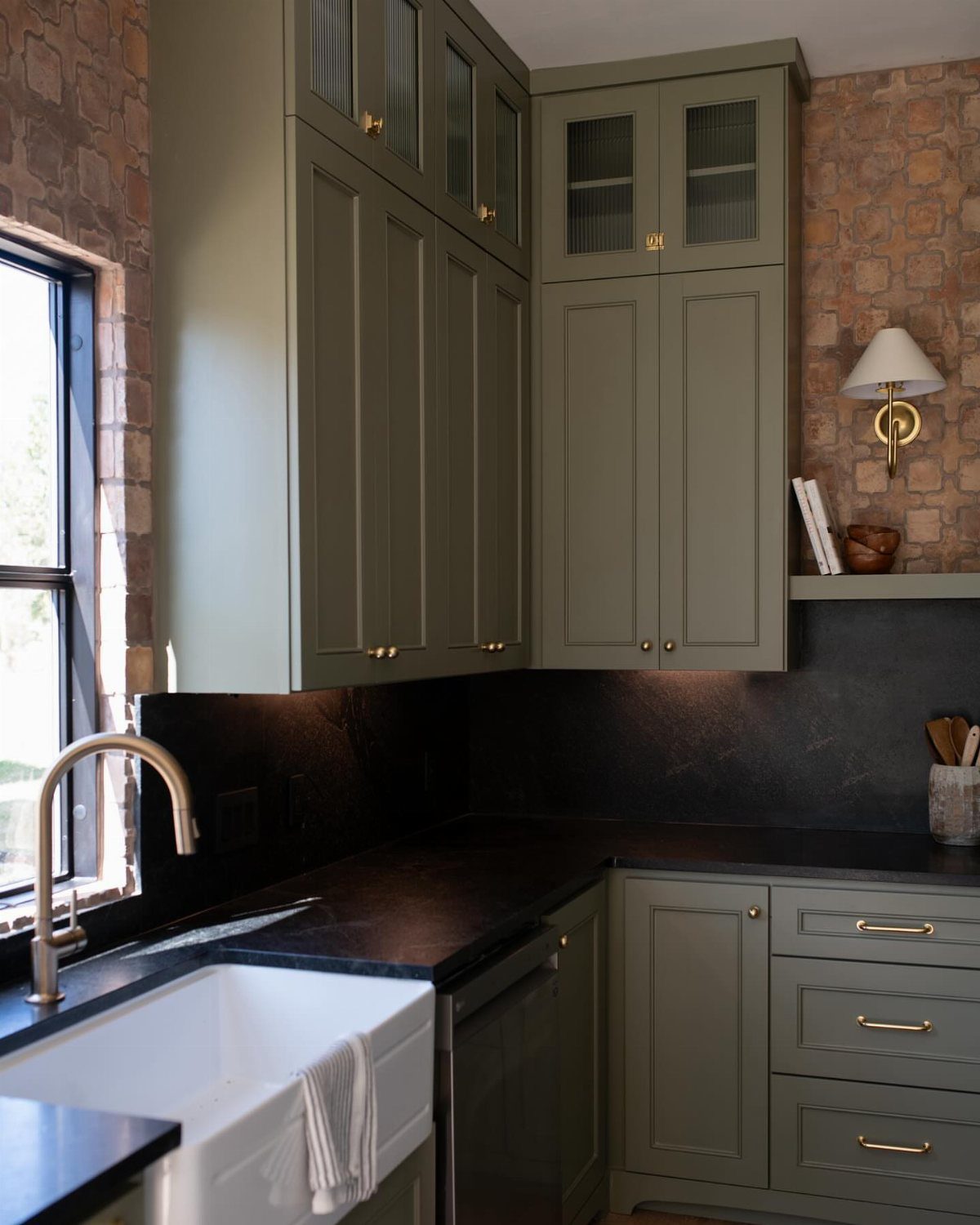 Olive green kitchen cabinets with black countertops, gold hardware, and exposed brick walls.