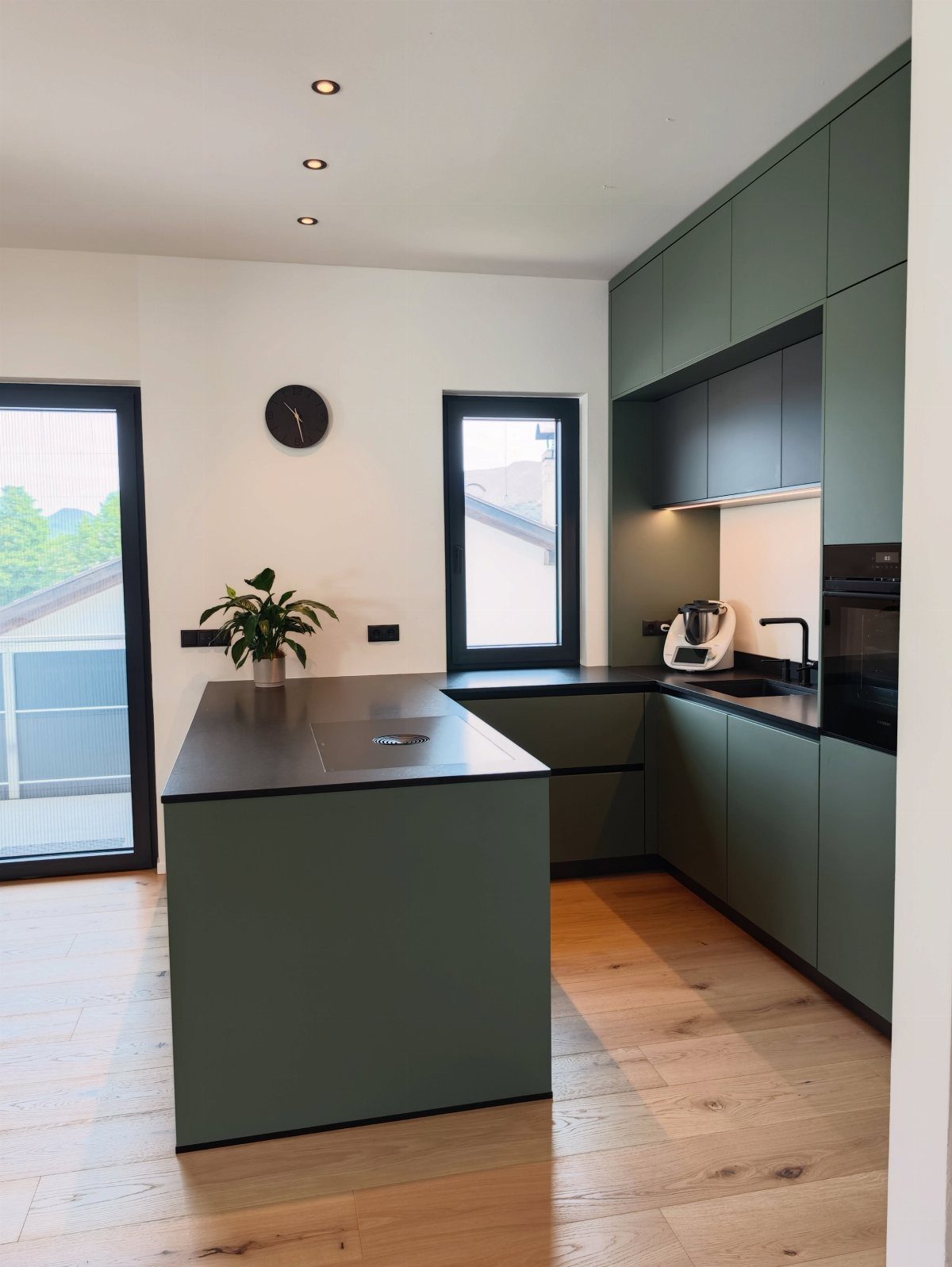 Modern kitchen with matte green cabinets, black countertops, and light wood flooring.