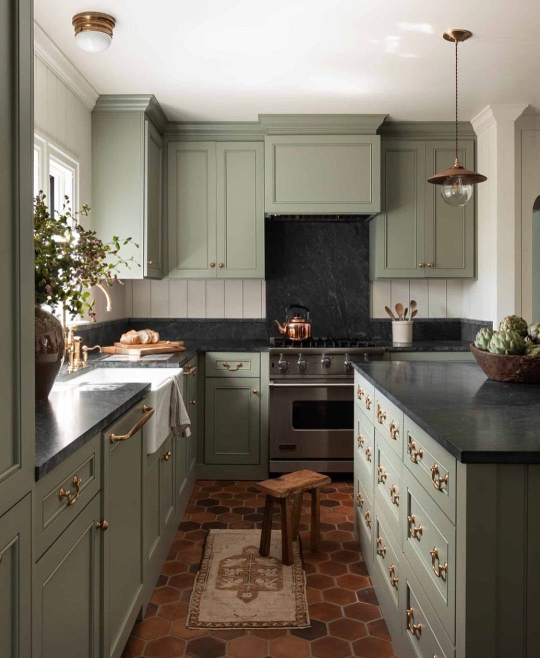 Sage green kitchen cabinets with black countertops and terracotta hexagonal floor tiles.