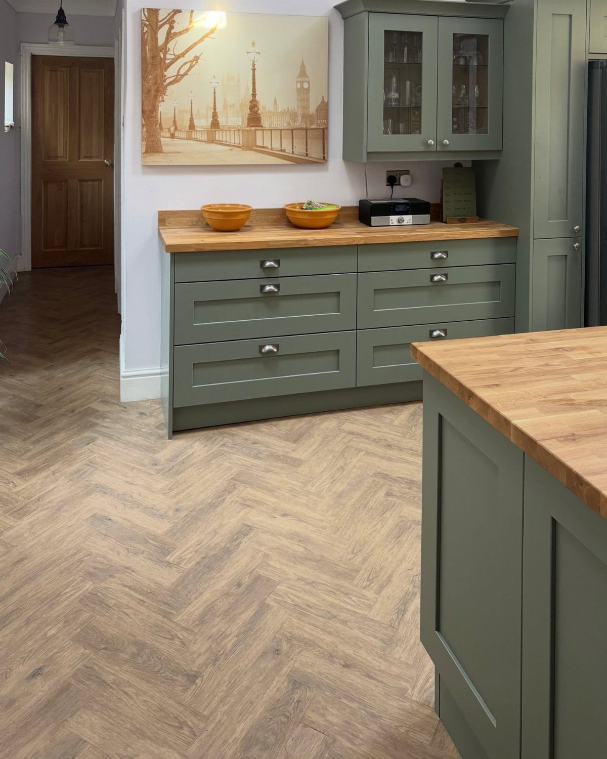Olive green kitchen cabinets with light wood butcher block countertops and herringbone flooring.