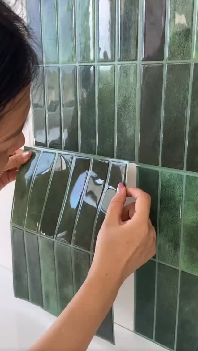 Hands applying a peel-and-stick sheet of glossy green subway tiles to a wall.