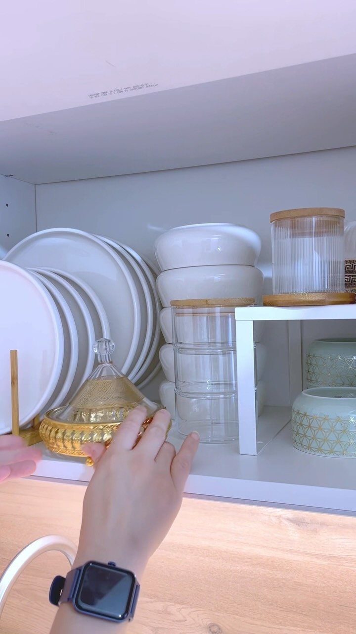 White kitchen cabinet with plates, bowls, and a white riser shelf holding jars.