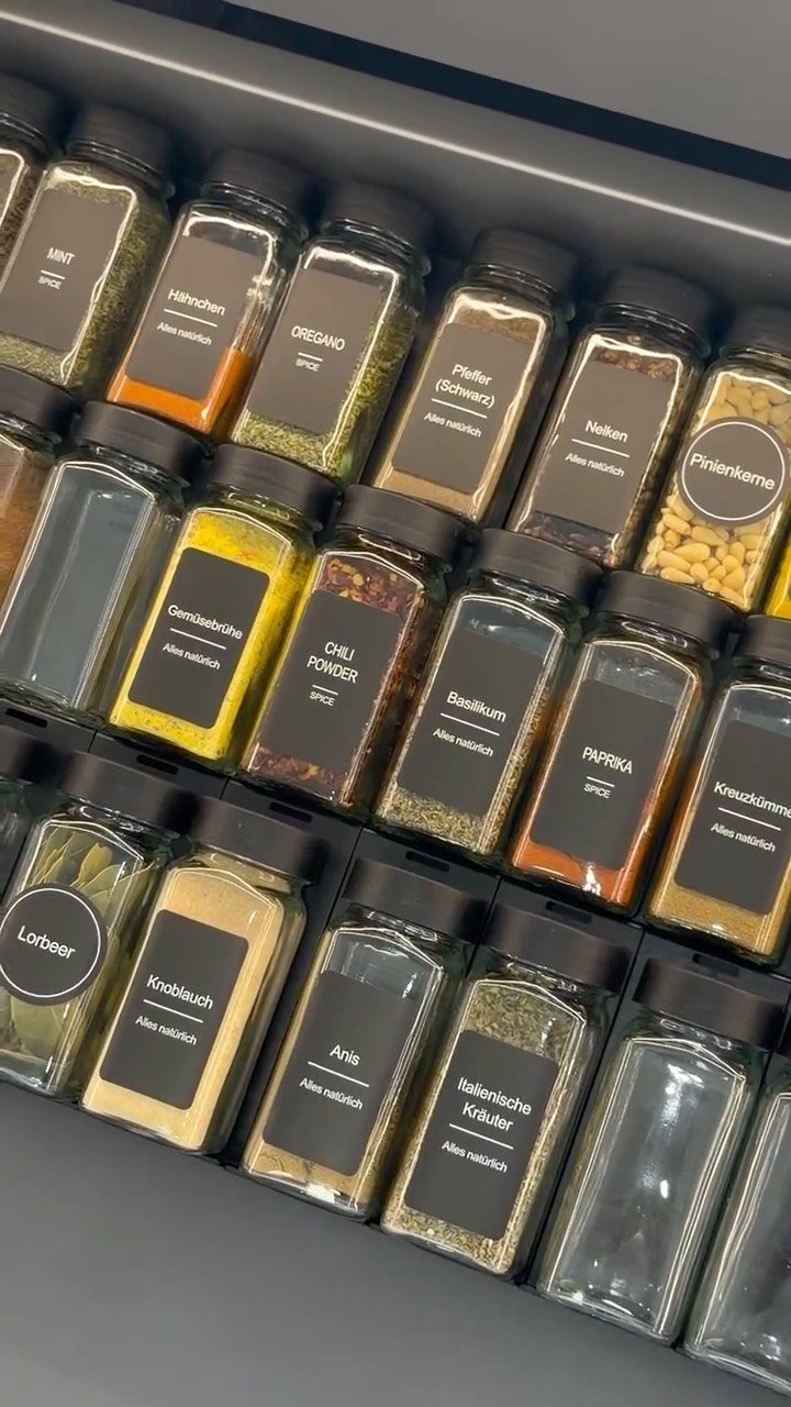 Clear glass spice jars with black labels organized in a dark grey drawer.