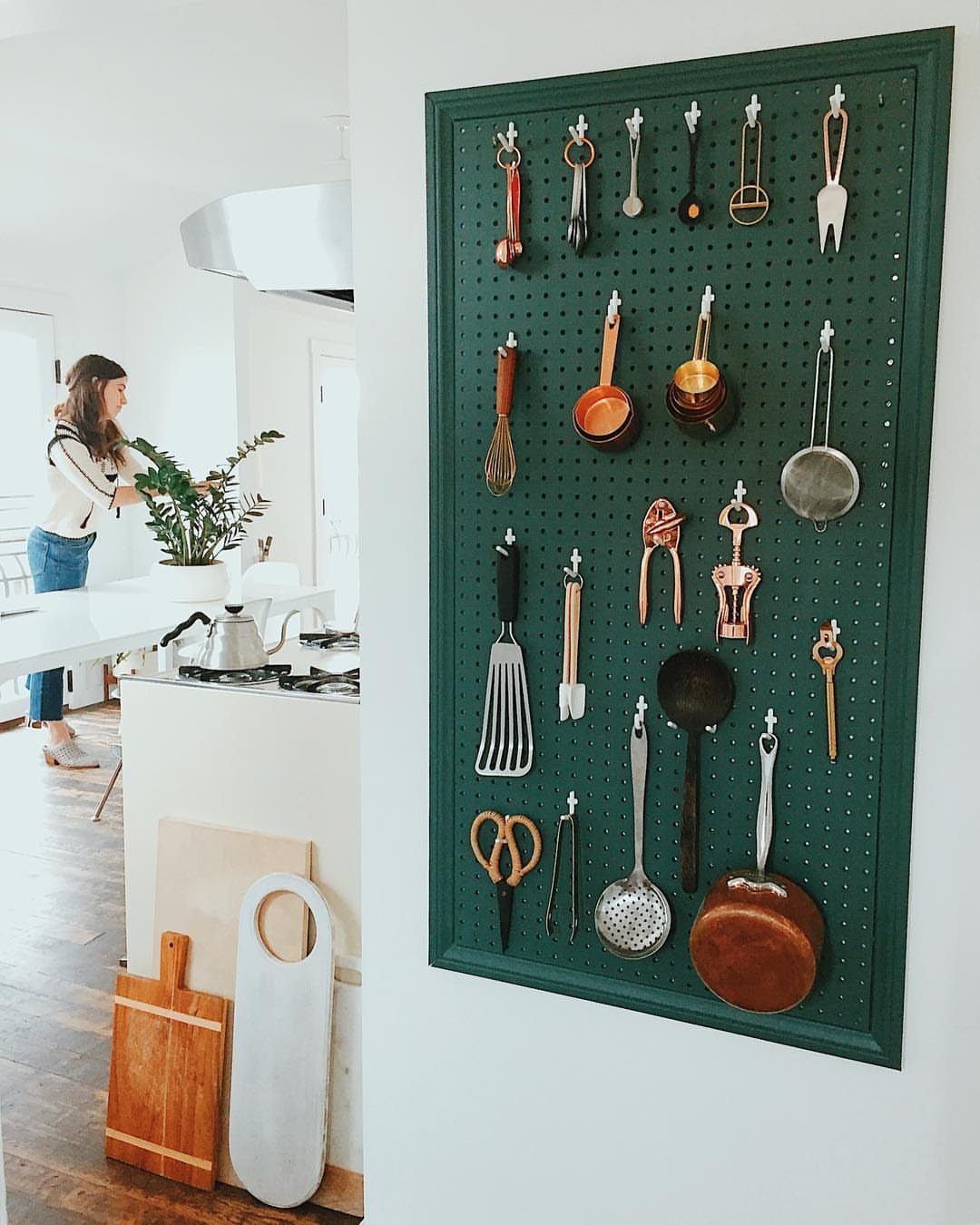 Green pegboard on a white wall holding various hanging kitchen utensils and pans.