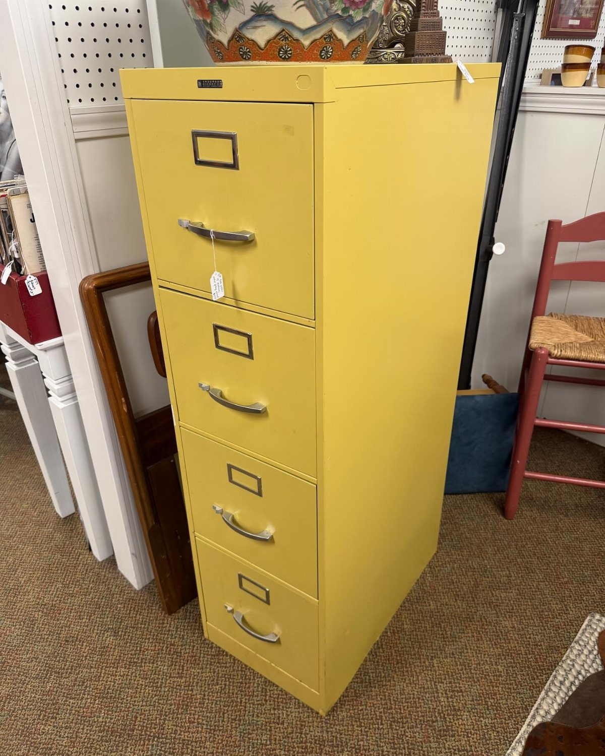 Bright yellow four-drawer metal filing cabinet standing on a brown carpeted floor.