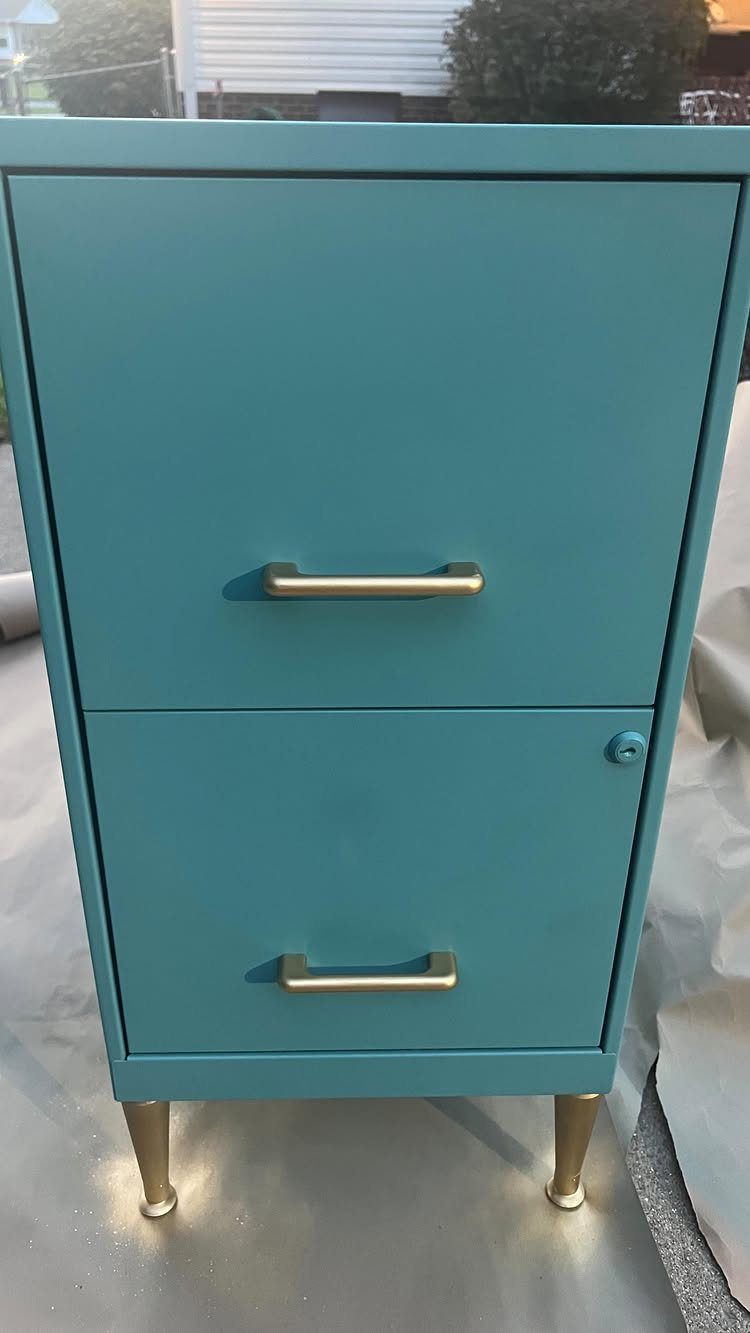 Teal metal file cabinet with gold handles and legs on a grey cloth.