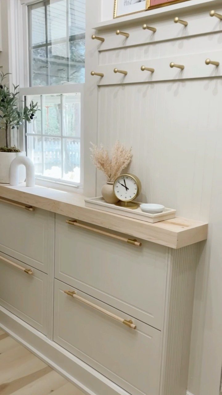 Light grey shoe cabinet with gold hardware and a wooden countertop in an entryway.