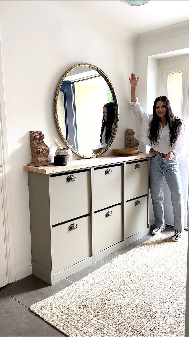 Light-grey shoe cabinet with wooden top and round gold mirror in a hallway.