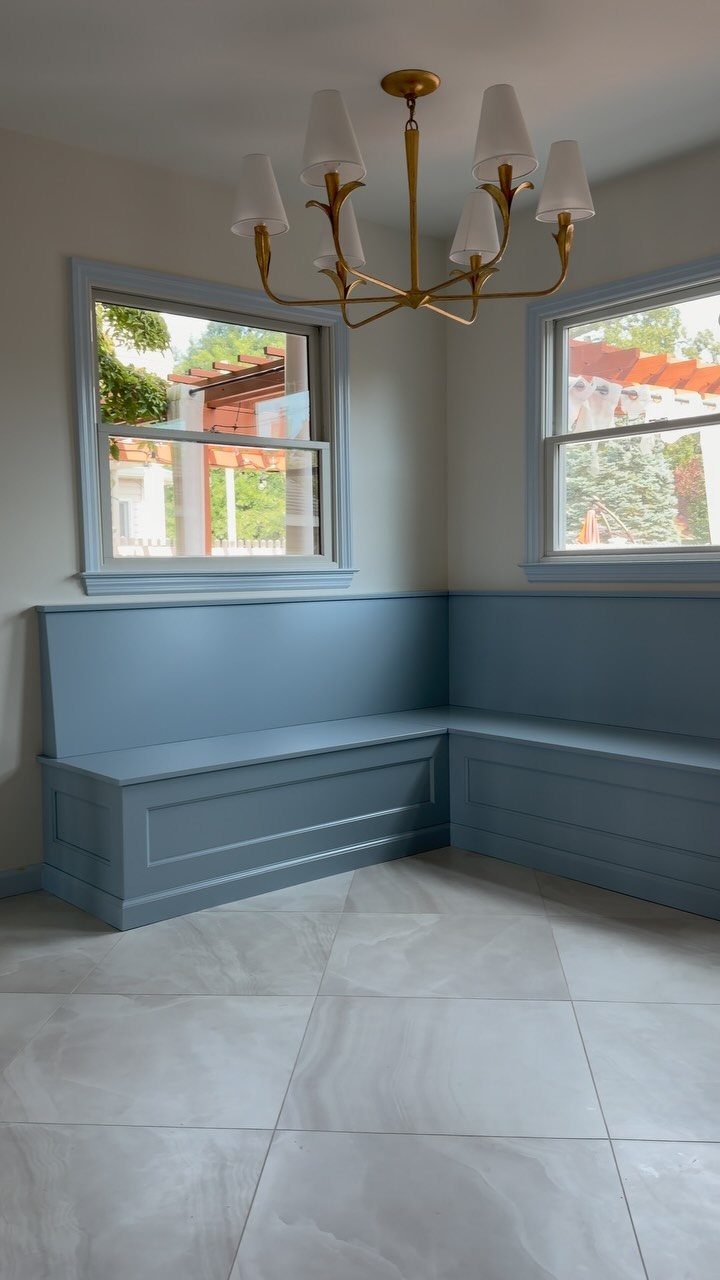 Blue L-shaped built-in bench in a corner under two windows with gold chandelier.