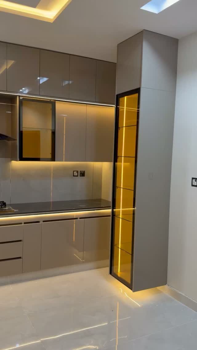 Glossy beige handleless kitchen cabinets with a tall illuminated glass display cabinet.