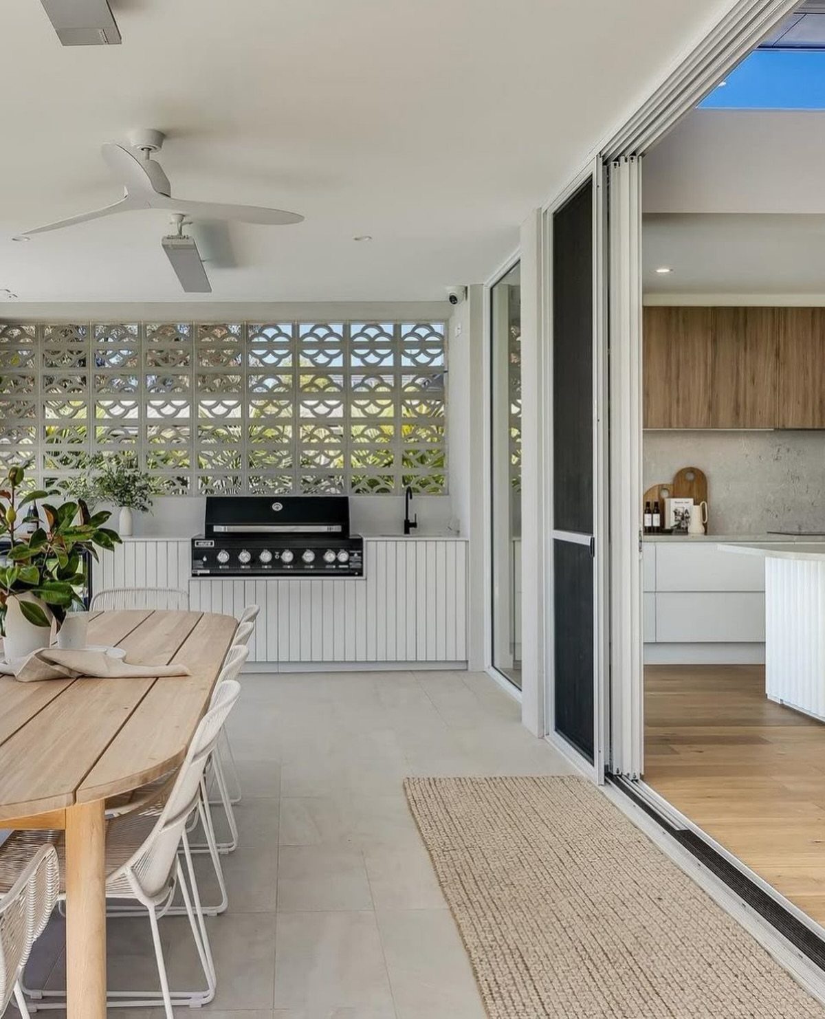 Outdoor dining area with wooden table and grill station opening into a modern kitchen