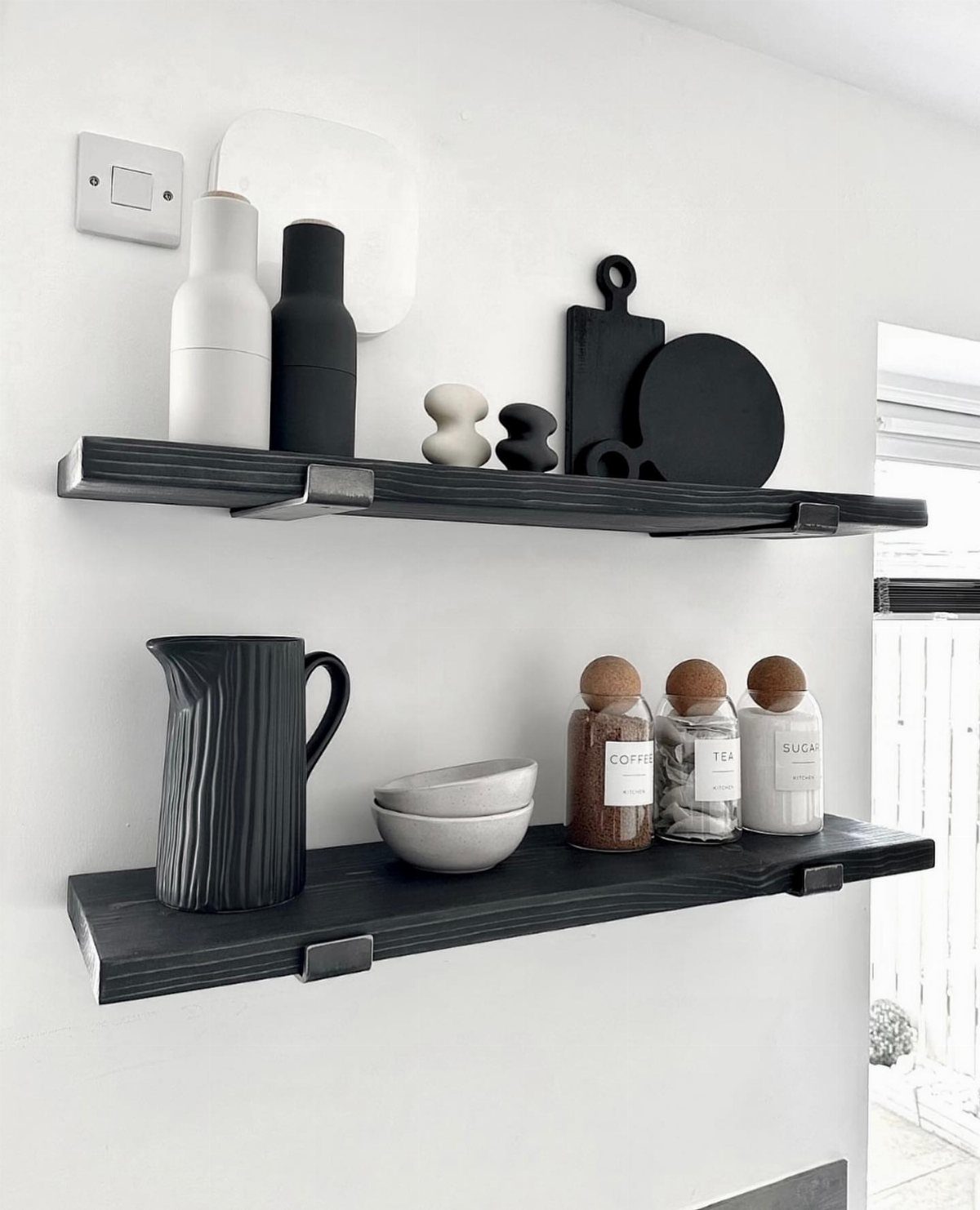 Black floating shelves on a white wall holding monochrome kitchen canisters and pitchers.