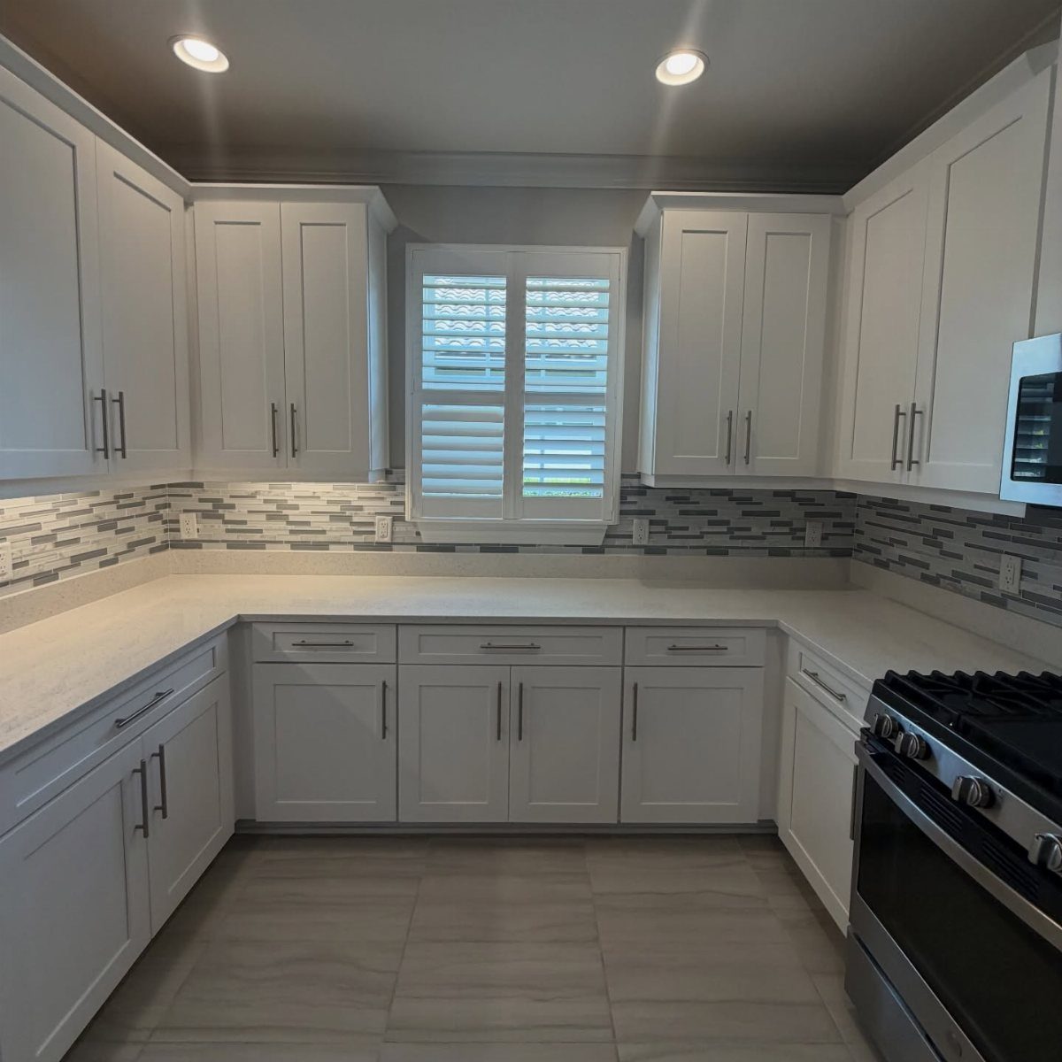White kitchen cabinets and countertops with a grey tiled backsplash and window shutters.