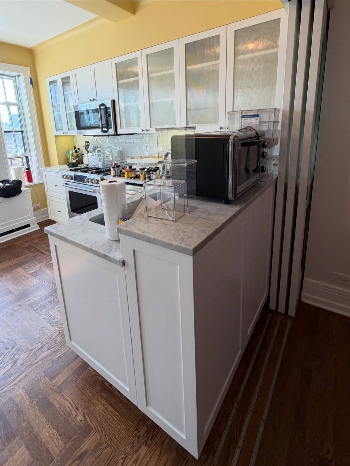 White kitchen cabinets and marble countertops against yellow walls with herringbone wood floors.