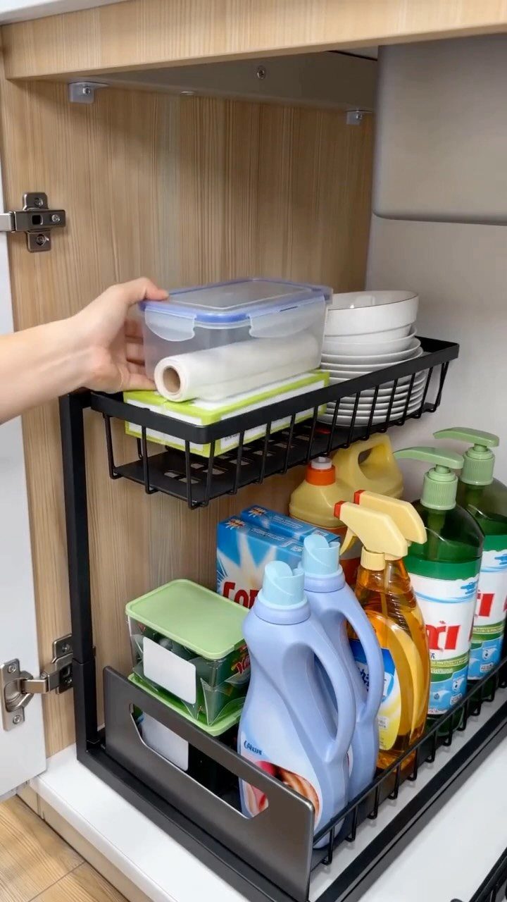 Black metal pull-out drawers in a light wood kitchen cabinet holding cleaning supplies.