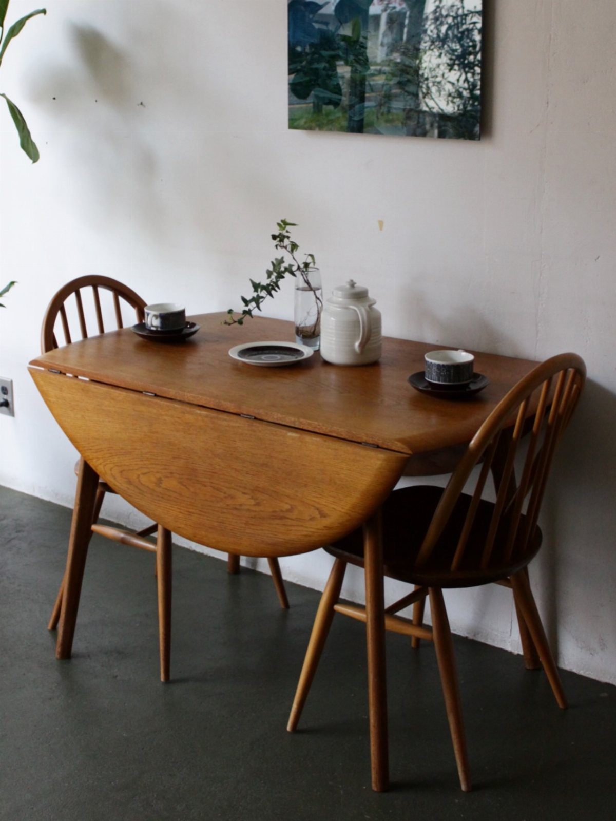 Wooden drop-leaf table with two matching chairs and coffee set against white wall.