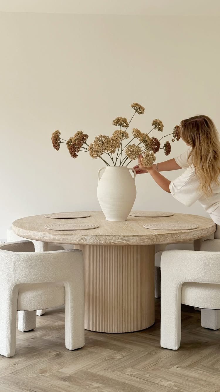 Round light-wood dining table with white textured chairs and a vase of dried flowers.
