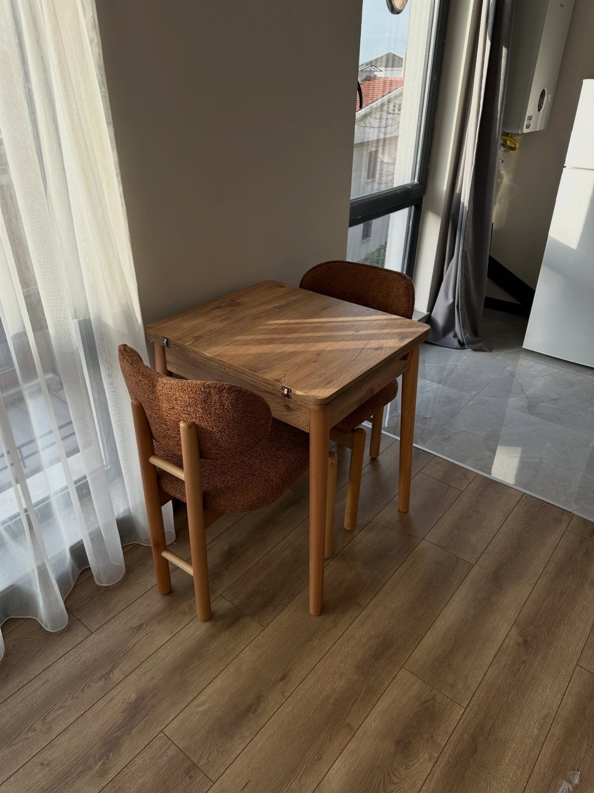Square wooden dining table with two brown chairs in a small bright corner.