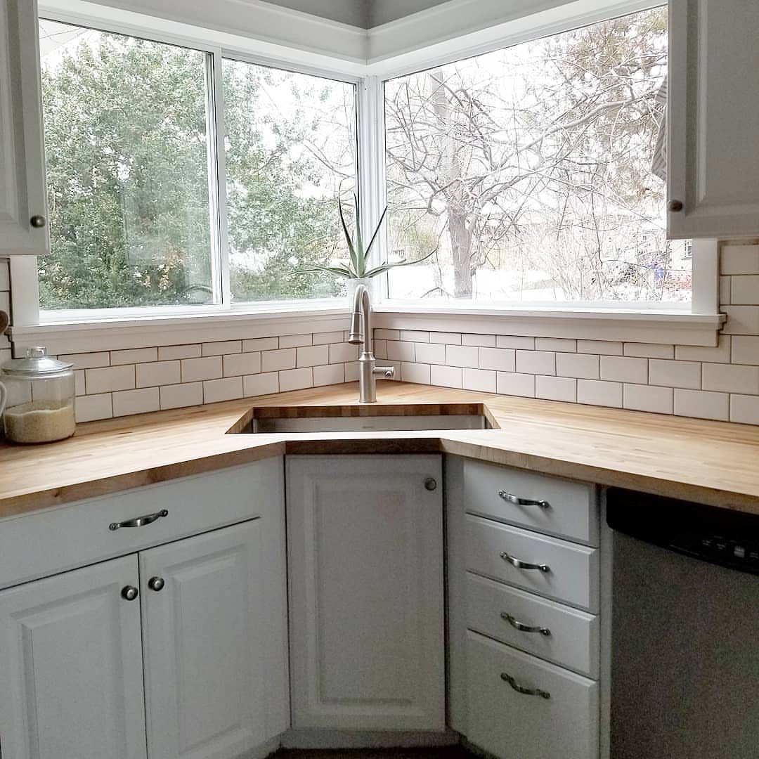 Corner sink built into a wooden countertop with white shaker cabinets and windows.