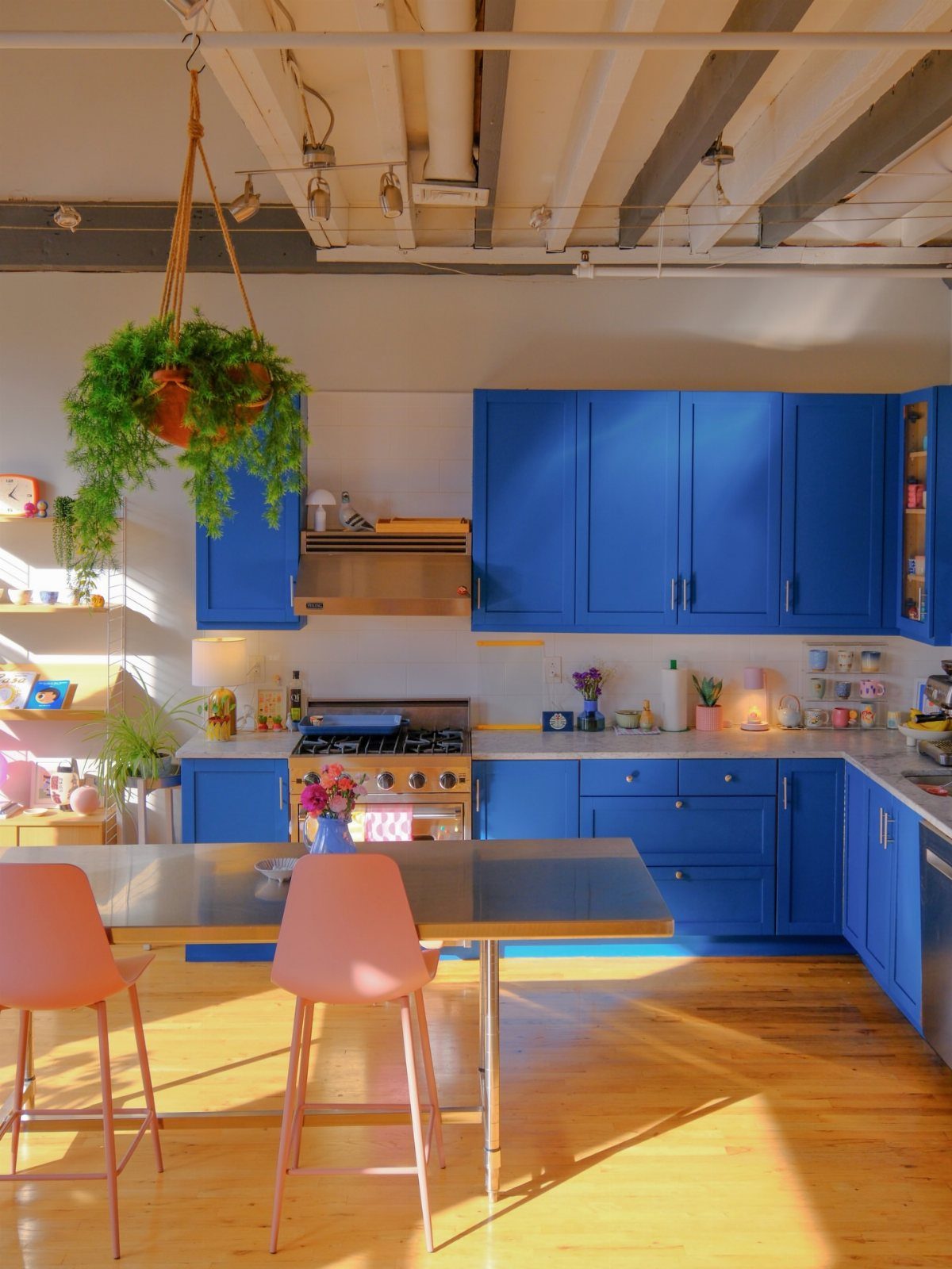 Blue kitchen cabinets with pink chairs and a hanging plant in industrial loft.