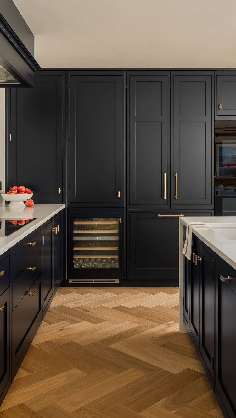 Matte black kitchen cabinets with gold hardware and light wood herringbone flooring.