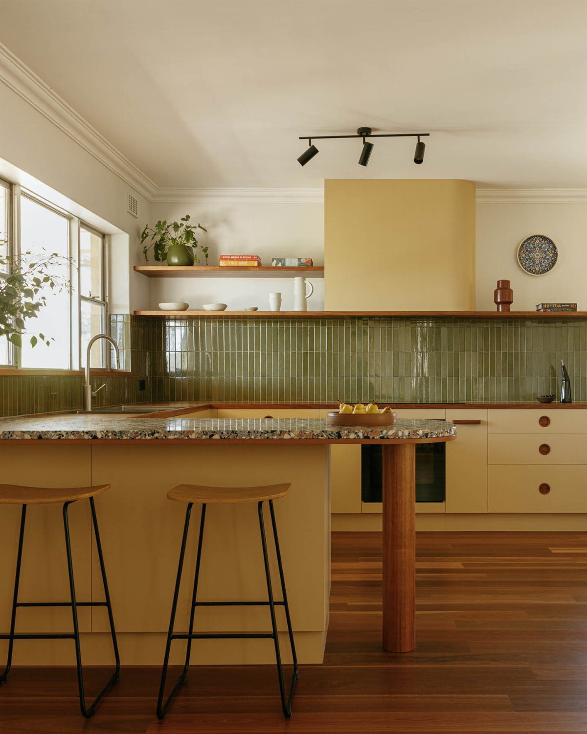 Kitchen island with two wooden stools, yellow cabinetry, and green tile backsplash.