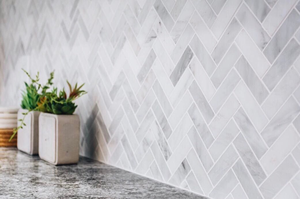 Marble herringbone mosaic backsplash with grey granite countertop and small potted plants.