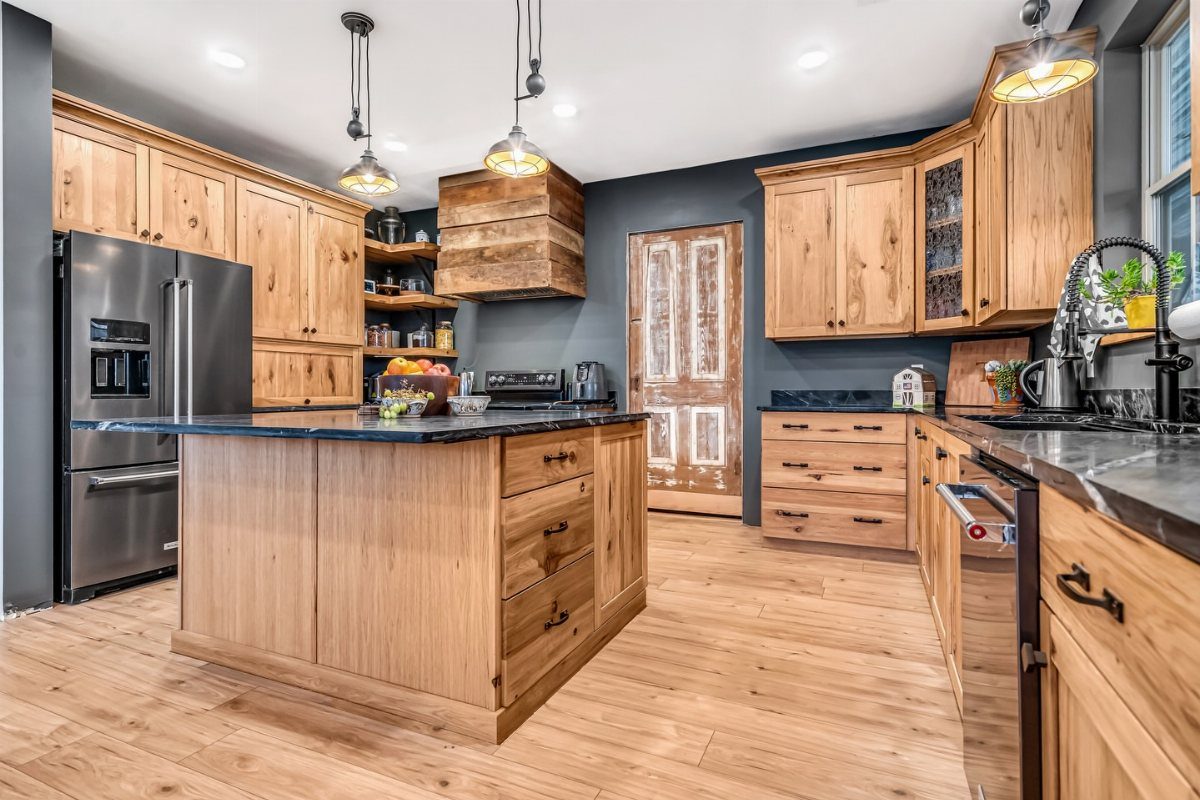 Natural wood kitchen cabinets and island with dark grey walls and black countertops.