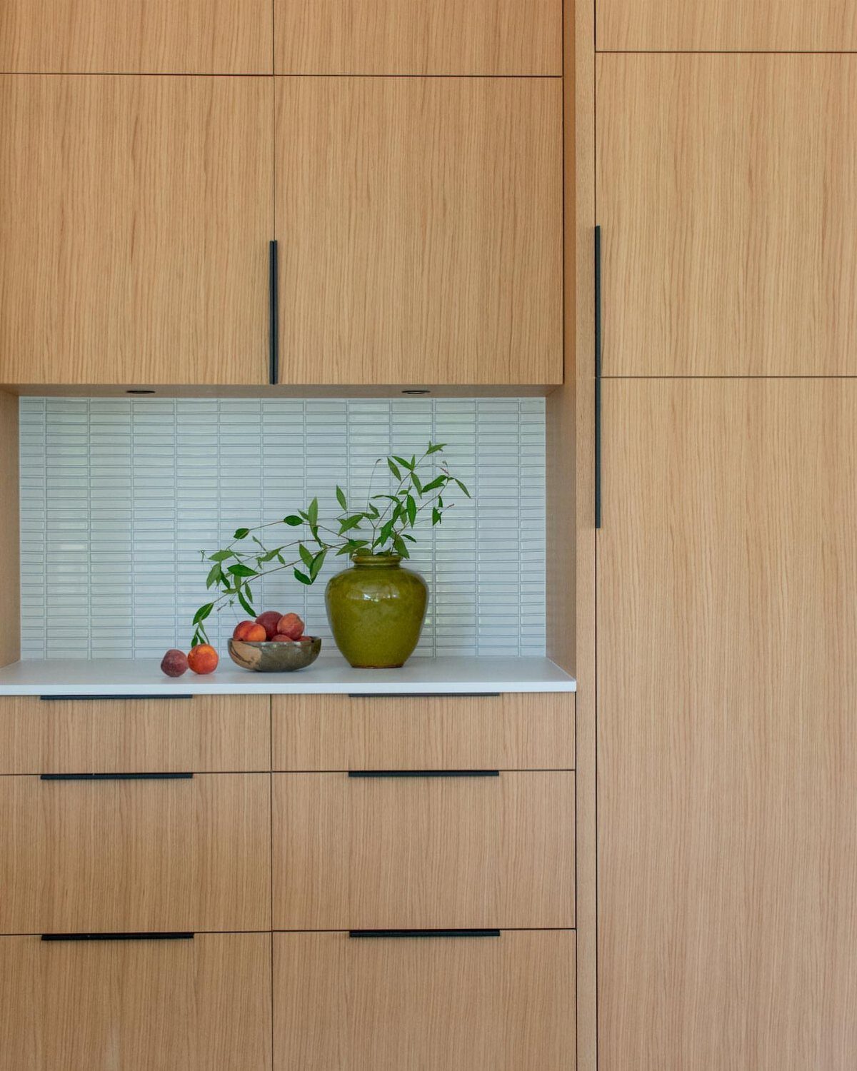 Light oak kitchen cabinets featuring modern black linear handles and white tiled backsplash.