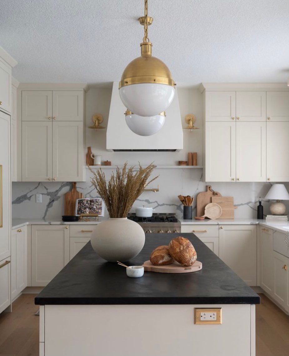Off-white kitchen cabinets with a black island countertop and gold pendant lighting.