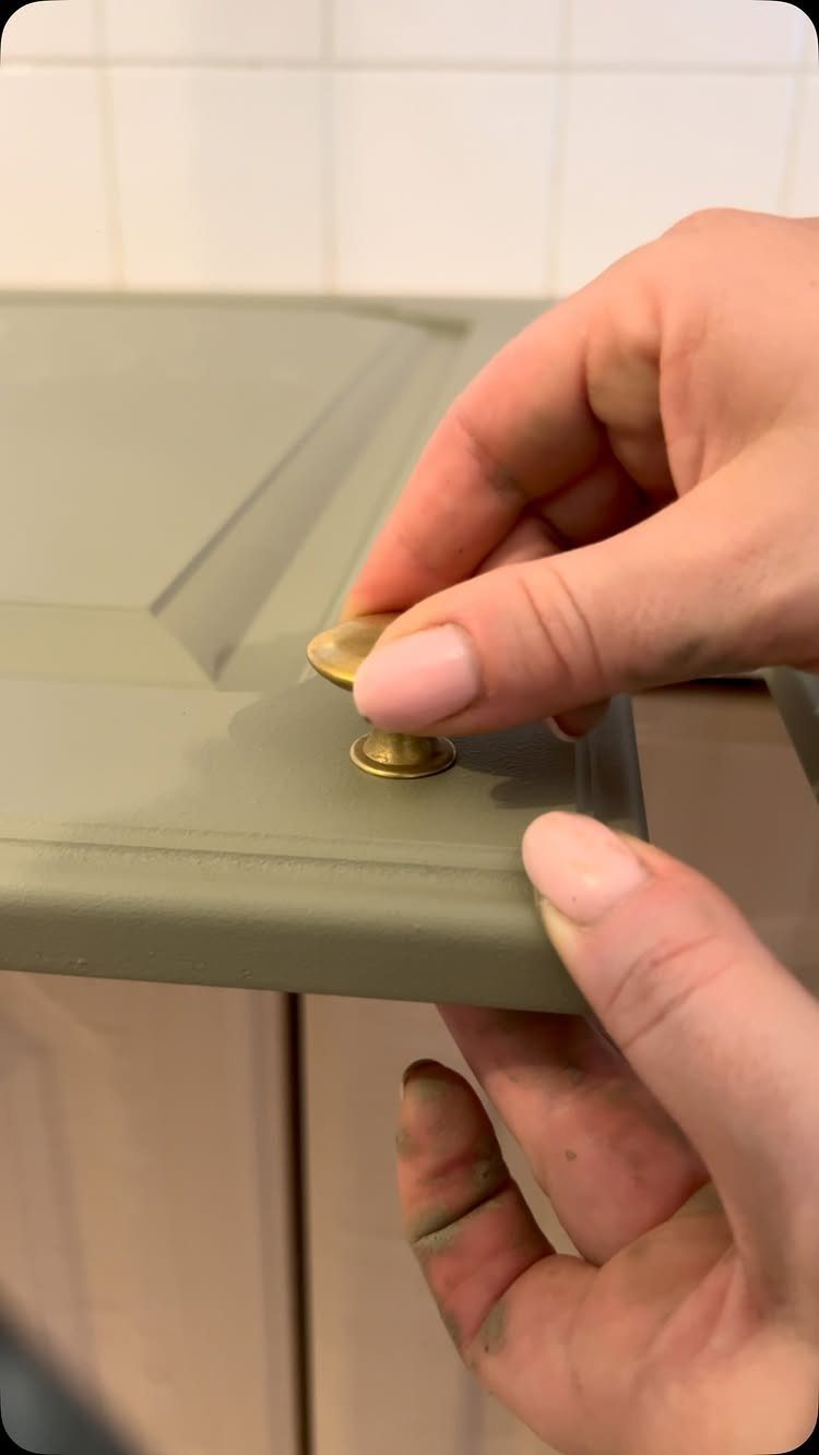 Hands installing a round brass knob on an olive green kitchen cabinet door.