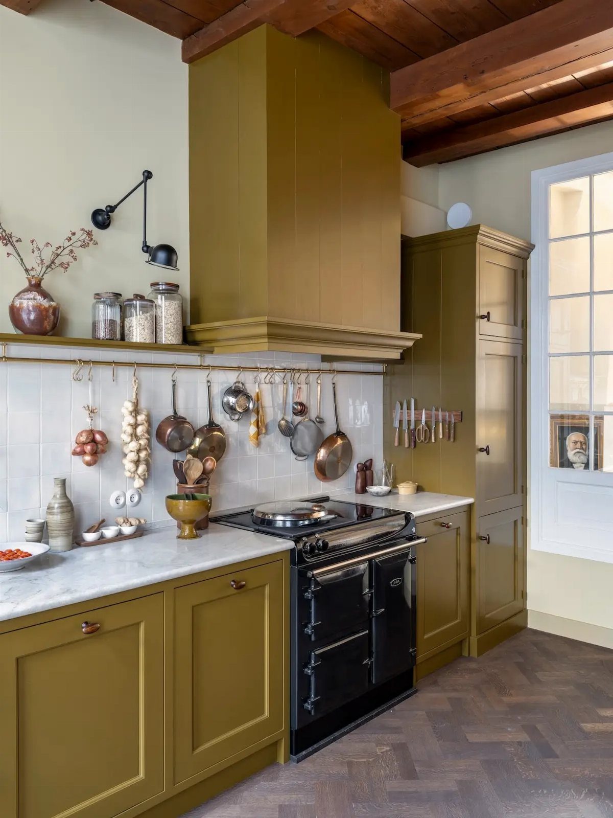 Olive green kitchen cabinets with white marble countertops and dark wood ceiling beams.