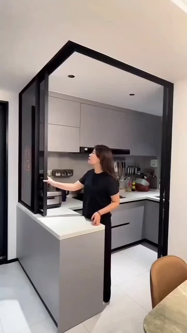 Woman gesturing toward black-framed glass sliding doors dividing a modern grey kitchen.