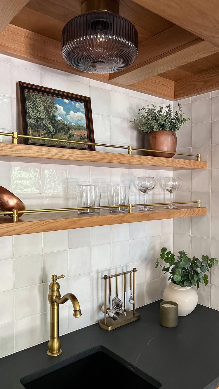 Wooden floating shelves with gold rails holding glassware and a plant in a kitchen.