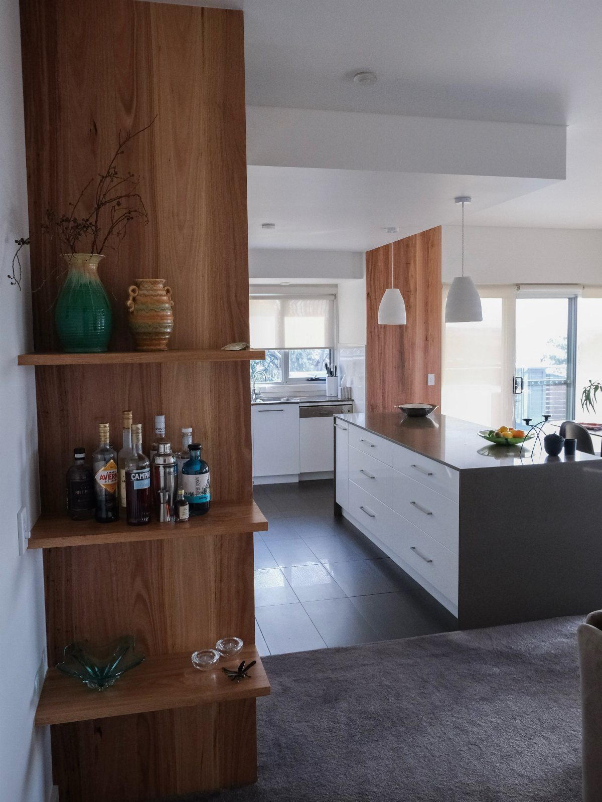 Wooden integrated kitchen shelving unit holding bottles and vases next to a modern island.