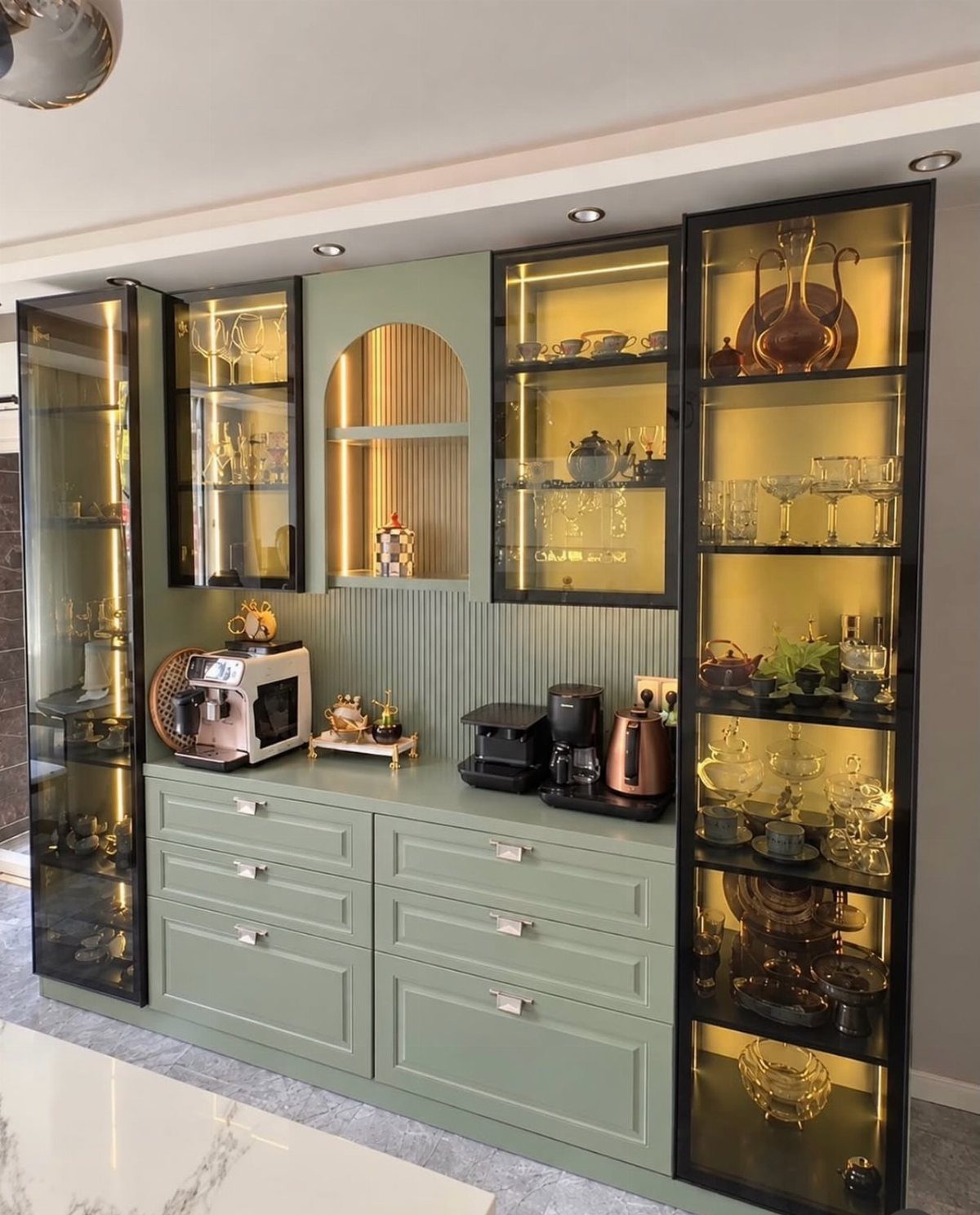 Sage green kitchen cabinets with illuminated glass-front upper displays and silver drawer handles.
