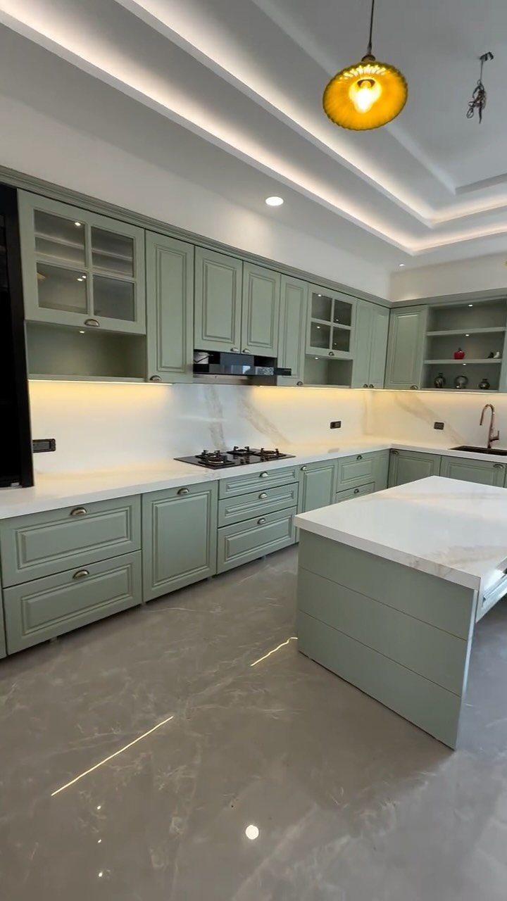 Sage green kitchen cabinets and island with white marble counters and gray floors.