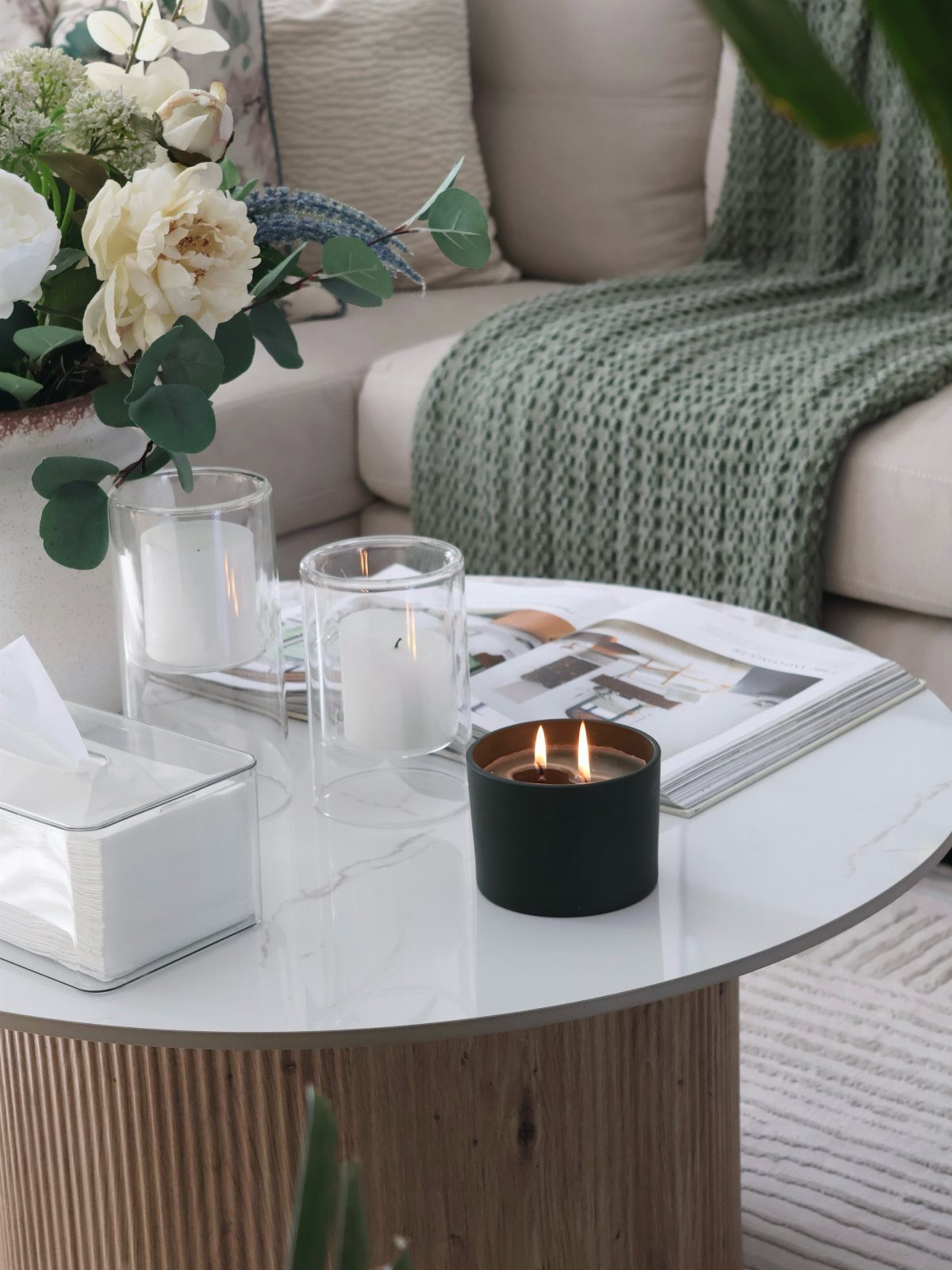 White marble round coffee table with candles, magazine, and flowers near a beige sofa