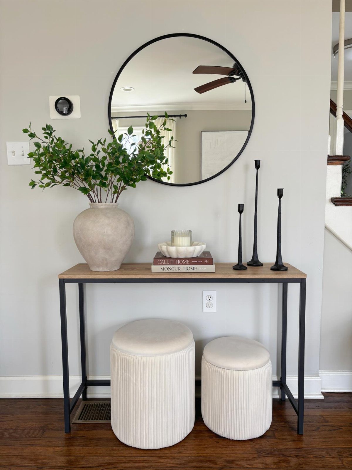Wooden console table with black legs, white ottomans, and a round wall mirror.