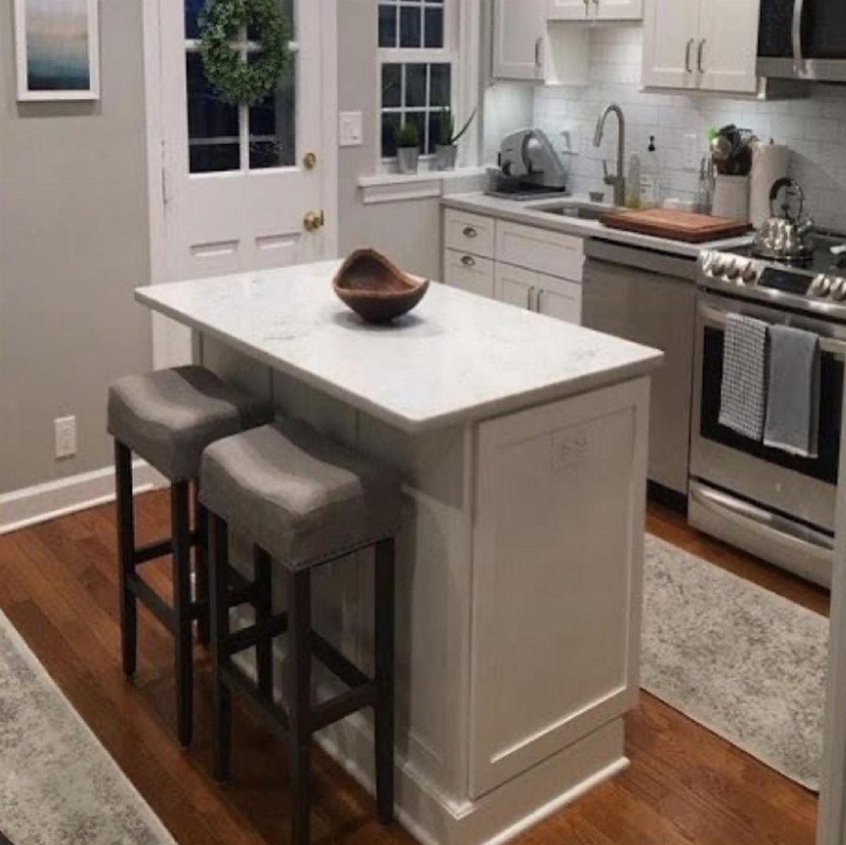 White kitchen island with marble countertop and two grey bar stools on hardwood floors.