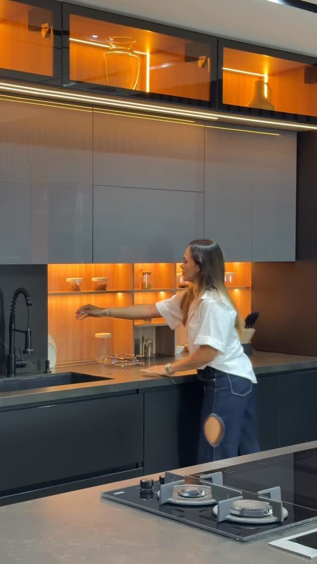 Woman standing in a modern kitchen with grey high-gloss cabinets and orange lighting.