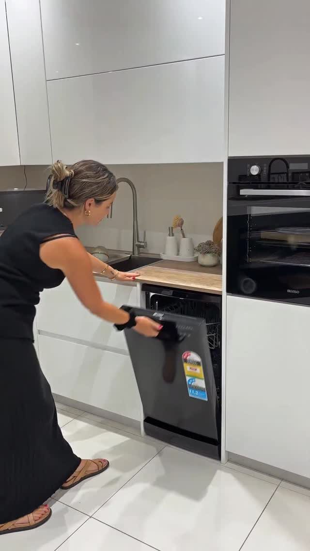 Woman opening a slim black dishwasher in a modern white kitchen setting.